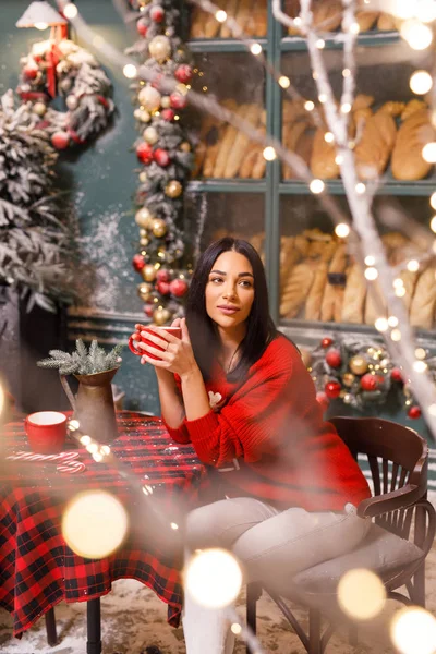 若い美しいブルネットの女の子赤いセーターと白いパンツパン屋の窓を背景にパリの路上でお茶を飲む — ストック写真
