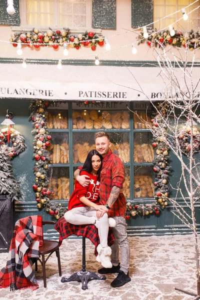 若い美しいカップル 赤いセーターと白いパンツのブルネットの女の子 赤いシャツの男はパン屋の窓の背景にパリの通りに座っています — ストック写真