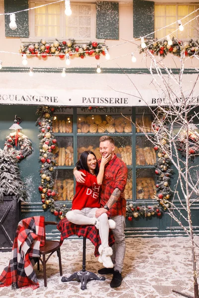 若い美しいカップル 赤いセーターと白いパンツのブルネットの女の子 赤いシャツの男はパン屋の窓の背景にパリの通りに座っています — ストック写真