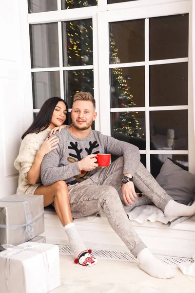 若い美しいカップルブルネットの女の子白いセーターにブロンドの男灰色のセーター彼のアパートの窓の近くに座って — ストック写真