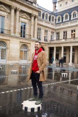 Paris 'te bir meydanda kahverengi ceketli ve kırmızı kazaklı genç bir adam duruyor. Fransa