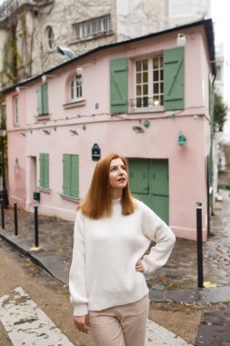 Beyaz kazak ve pembe pantolon giymiş güzel bir kız, Paris 'in güzel sokaklarında pembe bir evin arka planında yürüyor.