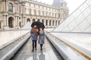 Gri montlu bir kız, gri montlu bir adam Paris 'teki Louvre Müzesi' nin yanında yürüyorlar.