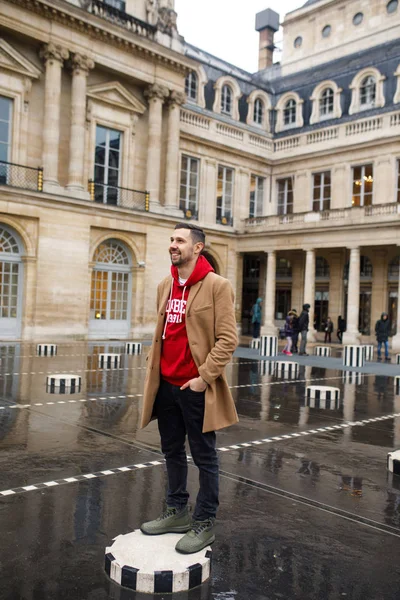 A young man in a brown coat and red sweater stands in a square in Paris. France