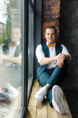 Çatı katındaki pencerenin yanında oturan klasik takım elbiseli genç adam.