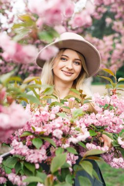 Beyaz elbiseli, gri pelerinli ve pembe şapkalı güzel sarışın kız Sakura ağaçlarının yanındaki parkta duruyor.