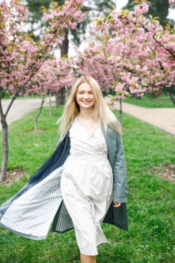 Gri pelerinli beyaz elbiseli güzel sarışın kız Sakura ağaçlarının yanındaki parkta duruyor.