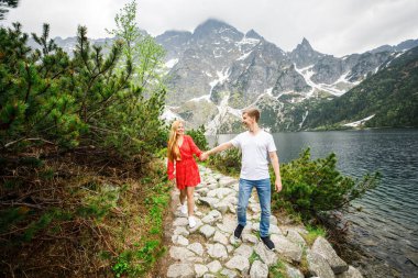 Genç bir çift, kırmızı elbiseli bir kız, beyaz tişörtlü ve mavi pantolonlu bir adam parktaki gölün yakınındaki bir dağın arka planında duruyor.