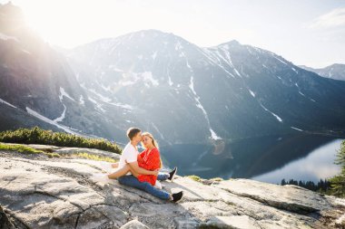 Genç bir çift, kırmızı elbiseli bir kız, beyaz tişörtlü ve mavi pantolonlu bir adam parktaki gölün yakınındaki bir dağın arka planında duruyor.