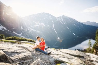 Genç bir çift, kırmızı elbiseli bir kız, beyaz tişörtlü ve mavi pantolonlu bir adam parktaki gölün yakınındaki bir dağın arka planında duruyor.