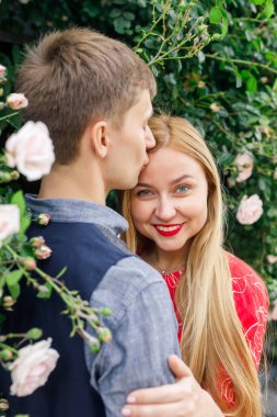 Genç bir çift şık giyiniyor, kırmızı elbiseli bir kız, mavi gömlekli ve mavi pantolonlu bir adam gül çalılığının yanında duruyor.