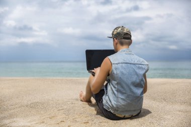 Genç adam sahilde otururken dizüstü bilgisayarda çalışıyor. Serbest çalışma, sosyal ağlar kavramı