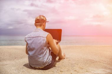Genç sahilde otururken dizüstü bilgisayar üzerinde çalışan adam. Serbest çalışma, sosyal ağlar kavramı. Tonda