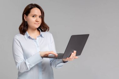 Serbest çalışan çekici genç bir kadın, ofisten uzak, dizüstü bilgisayarını tut, üniversite kompozisyonunu yaz, sola dön gülümseyen fotokopi alanına dön, gri arka planda dur, bilgisayarla interneti tara.