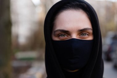 Yüz maskesi takan genç bir kadın. Siyah kapüşonlu yakışıklı kadın siyah tıbbi maske takıyor. Portreyi kapat. Salgın koronavirüs covid-19 karantina dönemi konsepti.