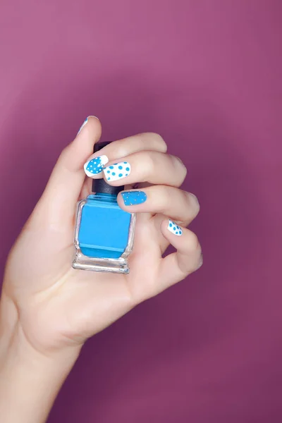 Hermosa manicura azul en las uñas en la mano con una botella de esmalte de uñas sobre un fondo rosa —  Fotos de Stock