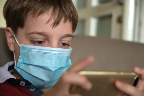 Little 9 year old boy in home isolation because of Covid 10 virus, wearing protective mask and playing video games on his cellphone.