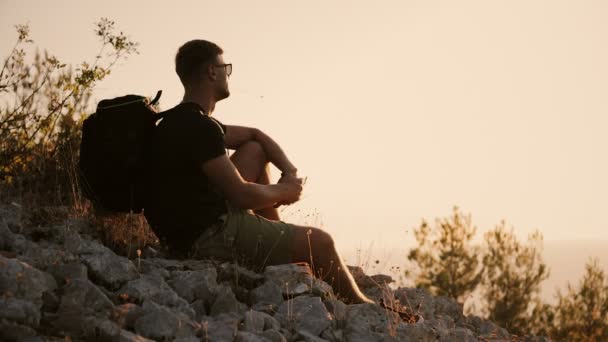 Um homem com uma mochila grande está sentado na montanha ao pôr do sol — Vídeo de Stock