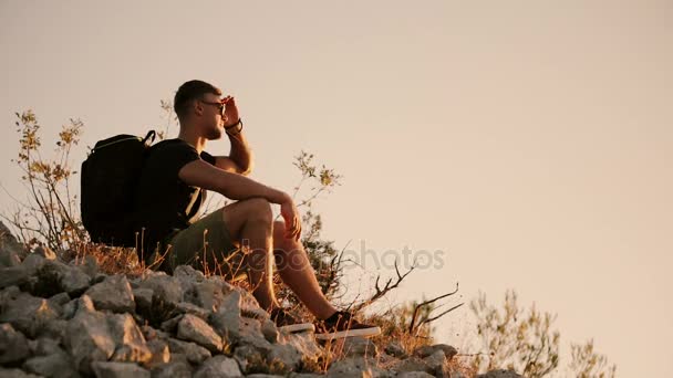 Ένας τουρίστας με ένα μεγάλο σακίδιο κάθεται στους βράχους και αναπαύεται στο ηλιοβασίλεμα — Αρχείο Βίντεο