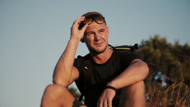 Slowmotion of Handsome young fashion man wearing sunglasses and backpack looks sunset in the field. 1920x1080 closeup — Stock Video