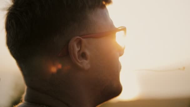 Slowmotion of Handsome young fashion man wearing sunglasses and backpack looks sunset in the field. In the mouth of a blade of grass. 1920x1080 closeup — Stock Video