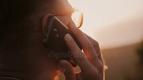 Un hombre con dos gafas de sol hablando en un teléfono móvil al atardecer . — Vídeo de stock