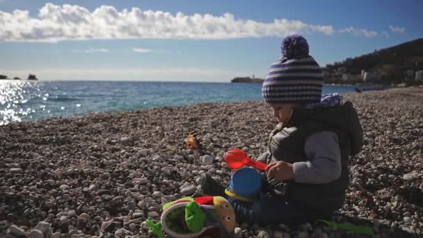 Un niño pequeño juega con juguetes en la orilla del mar con ropa de invierno y un sombrero . — Vídeos de Stock