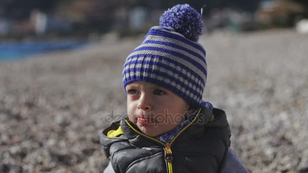 Un niño pequeño sonríe a la cámara y juega en la playa con guijarros . — Vídeos de Stock