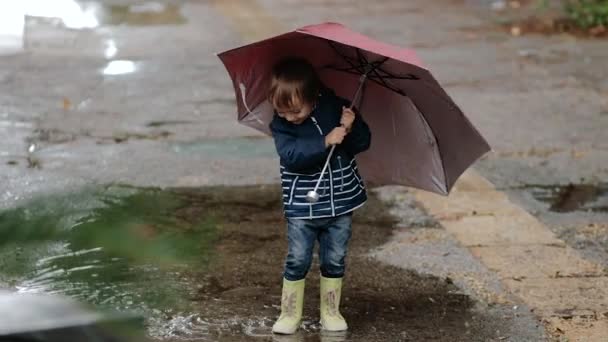 Chlapec stojí s deštníkem a gumovými botami v kaluži v dešti. — Stock video