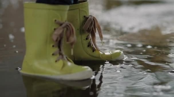 Un niño con botas de goma se para en un charco de agua bajo la lluvia de sus pies de cerca — Vídeo de stock