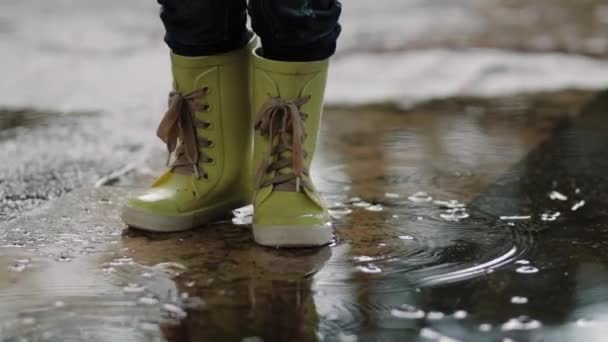 一个穿着橡胶靴的孩子站在水坑里，在雨中他的脚靠得很近 — 图库视频影像