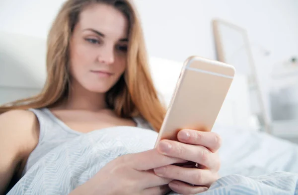 Jeune femme vérifiant son smartphone couché au lit — Photo