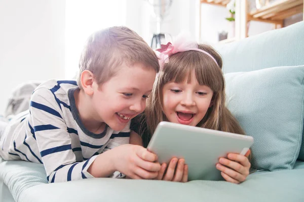 Irmãos felizes deitados no sofá em casa e brincando com tablet juntos — Fotografia de Stock