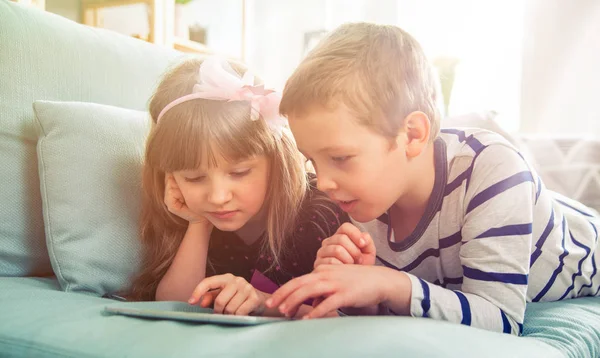 Irmãos felizes deitados no sofá em casa e brincando com tablet juntos — Fotografia de Stock