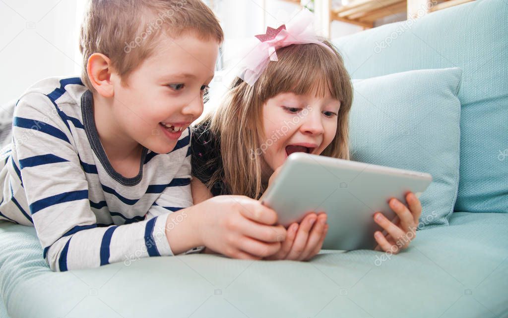 Happy siblings lying on sofa at home and playing with tablet together