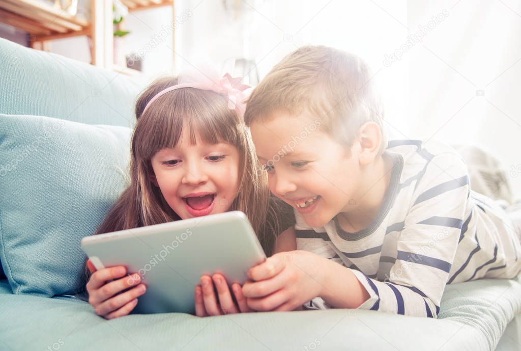 Happy siblings lying on sofa at home and playing with tablet together