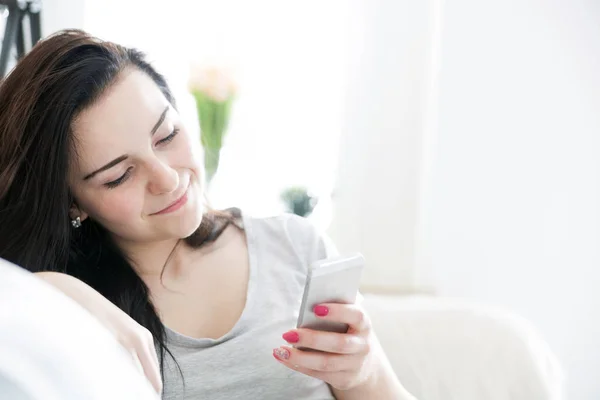 Junge Frau benutzt Handy, während sie zu Hause auf bequemem Sofa sitzt — Stockfoto