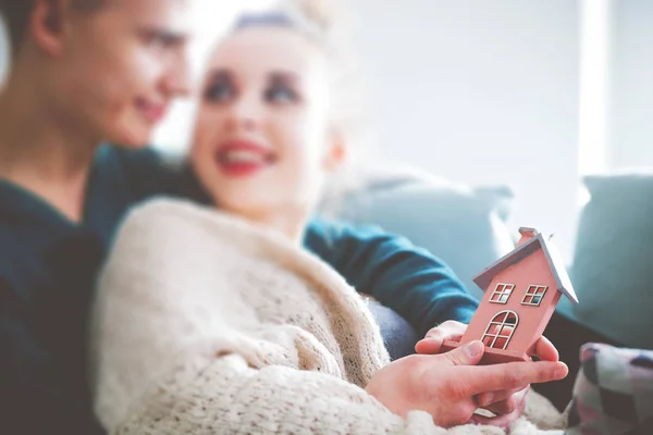 Amare la giovane coppia che si abbraccia sul divano di casa tenendo piccola casa in legno — Foto Stock