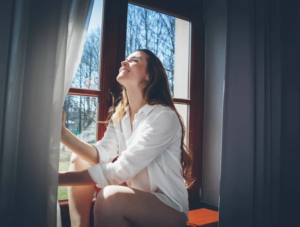 Mujer bonita en apartamento moderno abriendo cortinas de ventana después de despertar —  Fotos de Stock