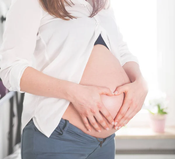 Dziewczyna w ciąży, trzymając się za ręce w kształcie serca na brzuch w domu — Zdjęcie stockowe