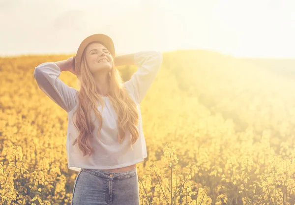 Mulher sorridente com chapéu no campo de colza amarelo ao pôr-do-sol — Fotografia de Stock