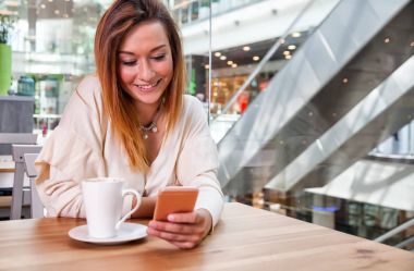 Rahat gülümseyen kız manifatura üstünde hareket eden telefon içinde café'de alışveriş merkezi