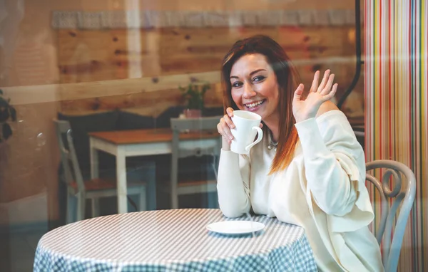 Ung flicka ler och vinkar till någon i café under dricka kaffe — Stockfoto