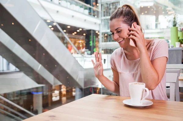 Girl before meeting with friend talking on phone and waving in cafe at shopping mall — Stock Photo, Image
