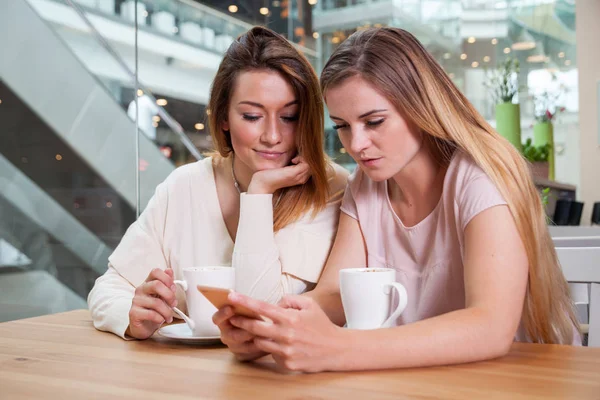 Dos amigas chicas en la cafetería del centro comercial usando el teléfono móvil y viendo contenido multimedia — Foto de Stock