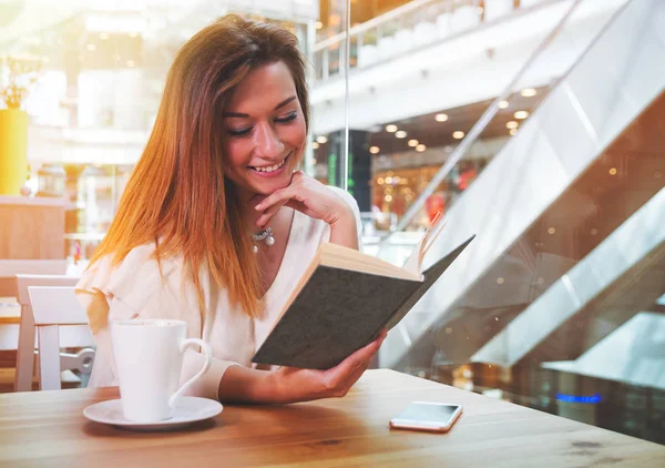 Расслабленная улыбающаяся девушка читает книгу в кафе в торговом центре — стоковое фото