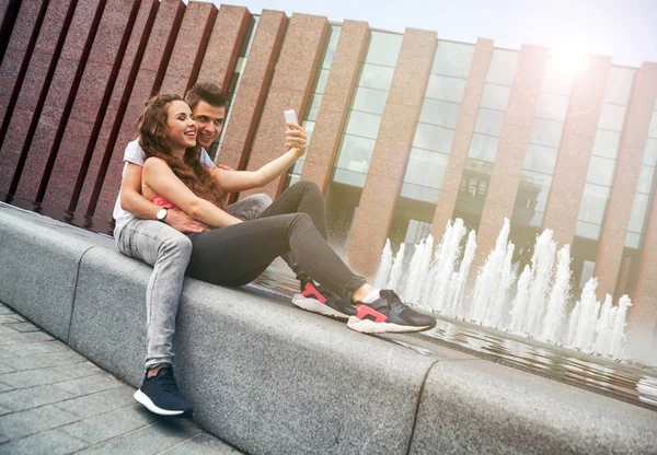 Pareja joven sentada en la ciudad al aire libre con teléfono móvil tomando se — Foto de Stock