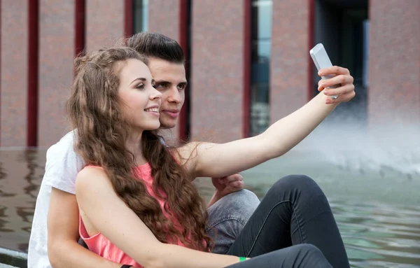 Młoda para siedzi w mieście odkryty z telefonu komórkowego biorąc selfie — Zdjęcie stockowe