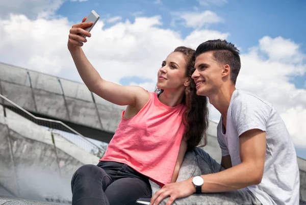 Młoda para siedzi w mieście odkryty z telefonu komórkowego biorąc selfie — Zdjęcie stockowe
