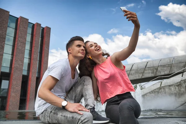 Młoda para siedzi w mieście odkryty z telefonu komórkowego biorąc selfie — Zdjęcie stockowe